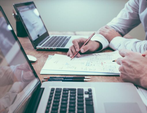 Duas pessoas conferindo papéis sobre uma mesa, com notebooks abertos ao lado